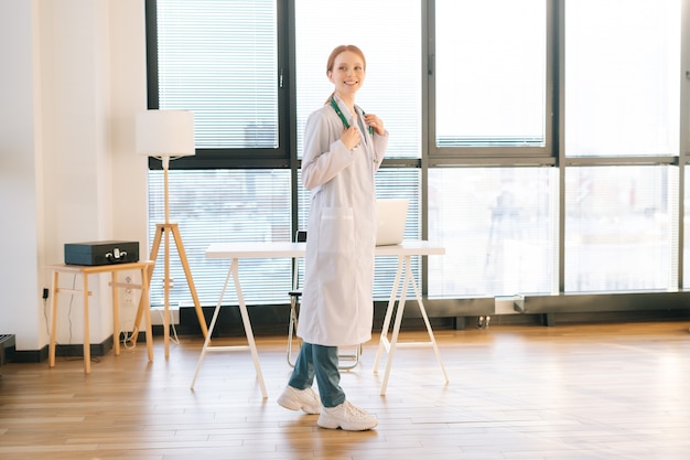 Vista frontal de la encantadora doctora en bata blanca de pie sobre el fondo de la ventana en un día soleado en la oficina de la clínica médica luz.