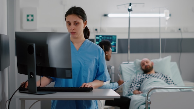 Vista frontal de la enfermera escribiendo experiencia médica en la computadora mientras en el fondo médico practicante negro discutiendo con el tratamiento médico del hombre enfermo. Paciente hospitalizado con trastorno respiratorio