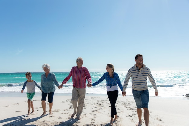 Vista frontal de una familia caucásica de varias generaciones en la playa con el cielo azul y el mar en el fondo, caminando, tomándose de la mano y sonriendo