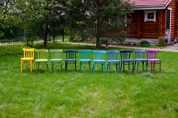 Vista frontal de una fila de sillas de madera multicolores sobre la hierba verde del jardín con una cabaña de troncos ...