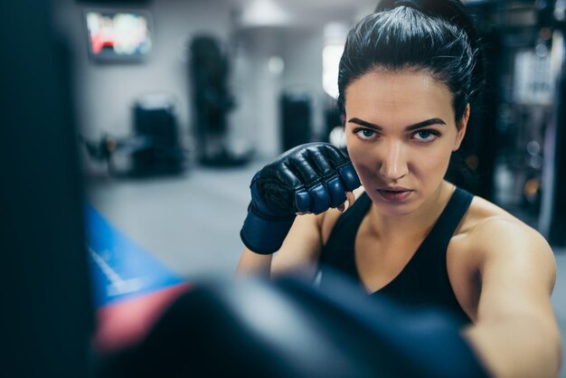 Vista frontal de una fuerte y atractiva entrenadora morena golpeando una bolsa con guantes de kickboxing en el entrenamiento del gimnasio Concepto de estilo de vida y motivación de fitness deportivo