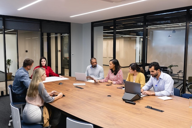 Vista frontal de un grupo multiétnico de hombres y mujeres sentados juntos junto a una mesa en la sala de reuniones, discutiendo e interactuando.