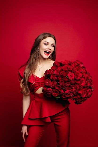 Vista frontal de la hermosa mujer en vestido rojo con maquillaje manteniendo un hermoso ramo de rosas mirando a la cámara y sonriendo Bonita modelo posando sobre fondo rojo aislado Concepto de día de San Valentín