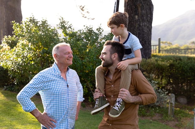 Foto vista frontal de un hombre caucásico en casa en su jardín, de pie con su hijo pequeño sentado sobre sus hombros, su padre mayor, de pie junto a ellos sonriendo con las manos en las caderas, el padre y así