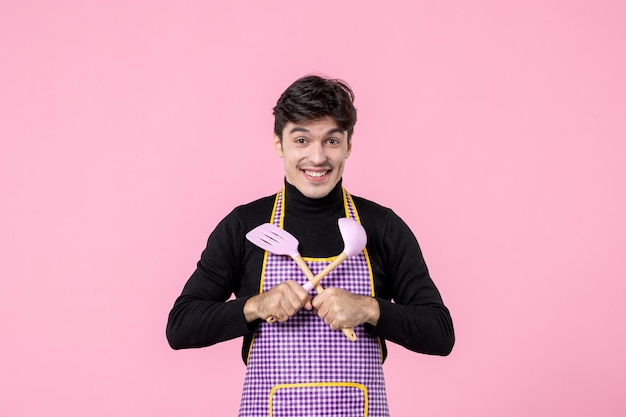 Vista frontal hombre joven en capa sosteniendo cucharas sobre fondo rosa profesión comida trabajo horizontal comida uniforme masa cocina color