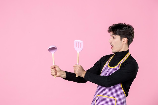 Vista frontal hombre joven en capa sosteniendo cucharas sobre fondo rosa trabajo masa profesión cocina uniforme horizontal cocina trabajador jefe colores