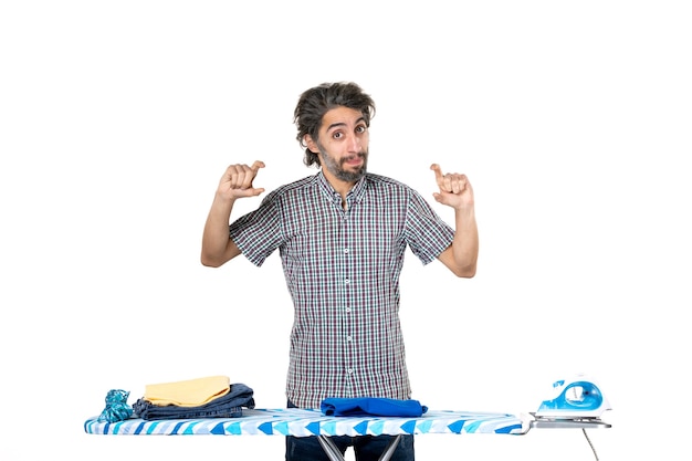 Vista frontal del hombre joven detrás de la tabla de planchar sobre fondo blanco máquina de trabajo de lavandería limpia hombre a casa quehaceres domésticos ropa