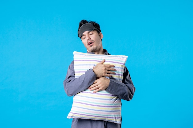 Vista frontal hombre joven en pijama sosteniendo la almohada y durmiendo sobre sus pies sobre fondo azul sueño humano dormir noche color tarde reposo en cama pesadilla despertar