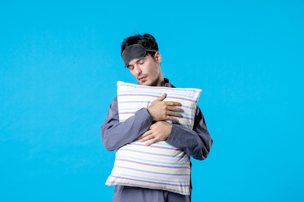 Vista frontal hombre joven en pijama sosteniendo una almohada sobre fondo azul sueño humano dormir noche color tarde reposo en cama pesadilla despertar cansado