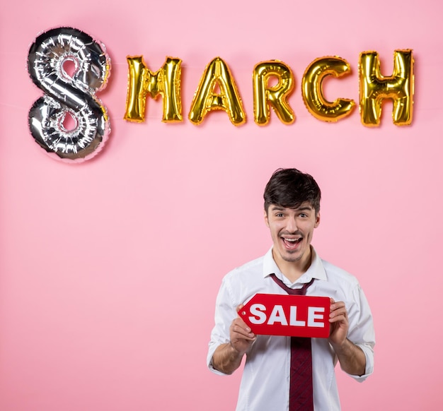 Vista frontal hombre joven con placa de identificación de venta roja con decoración de marzo sobre fondo rosa compras vacaciones color día de la mujer dinero fiesta femenina igualdad