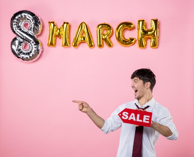 Vista frontal hombre joven con placa de identificación de venta roja con decoración de marzo sobre fondo rosa hombre femenino color fiesta compras presente igualdad vacaciones