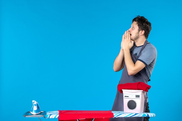 Vista frontal del hombre joven con tabla de planchar sobre la superficie azul