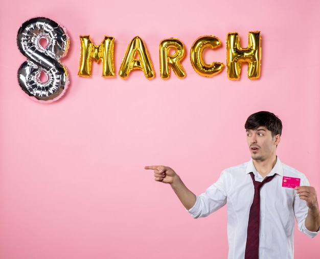 Vista frontal hombre joven con tarjeta bancaria rosa con decoración de marzo sobre fondo rosa hombre presente igualdad femenina compras dinero color vacaciones