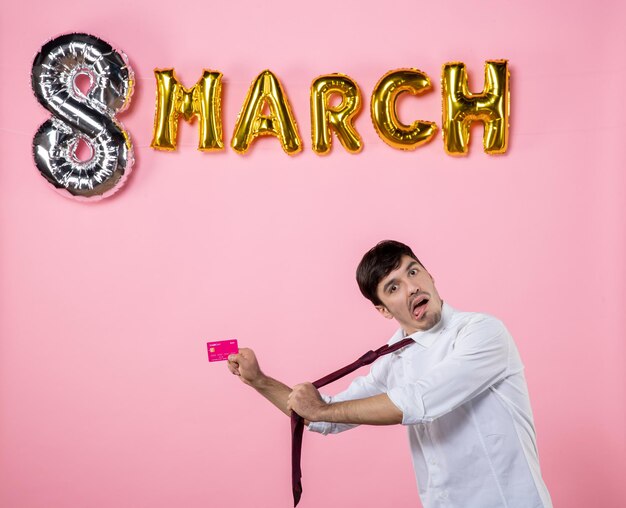 Vista frontal hombre joven con tarjeta bancaria rosa con decoración de marzo sobre fondo rosa igualdad femenina color dinero vacaciones compras hombre fiesta