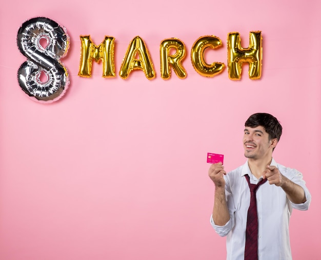 Vista frontal hombre joven con tarjeta bancaria rosa con decoración de marzo sobre fondo rosa presente igualdad femenina color vacaciones hombre fiesta dinero