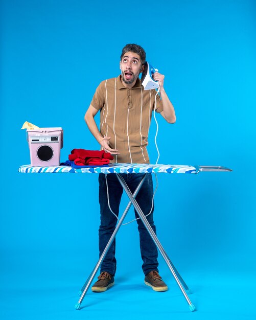 Foto vista frontal de los hombres jóvenes detrás de la tabla de planchar escuchando plancha sobre fondo azul color de las tareas del hogar limpiar a máquina lavado emoción hombre lavandería