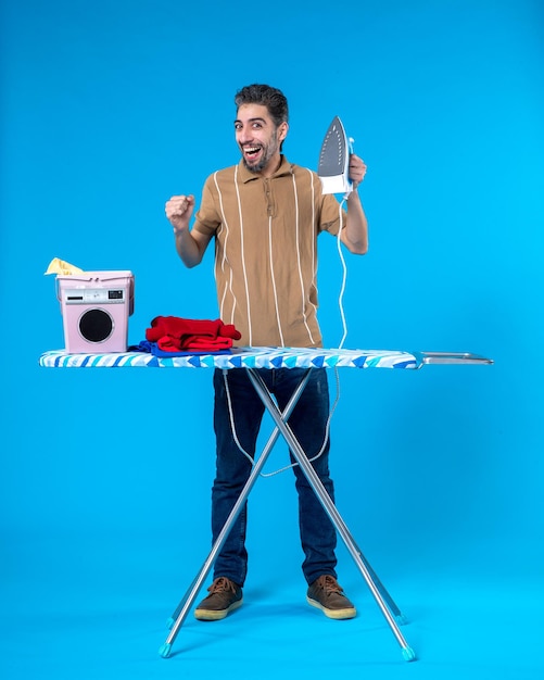 Vista frontal de los hombres jóvenes detrás de la tabla de planchar con plancha sobre fondo azul color de las tareas del hogar máquina limpia hombre lavandería emoción