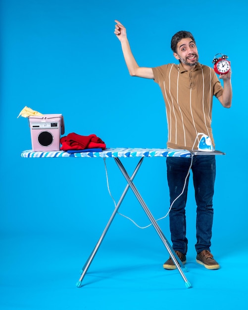 Vista frontal de los hombres jóvenes detrás de la tabla de planchar sosteniendo relojes sobre fondo azul color de las tareas del hogar lavadora tiempo de lavandería limpia