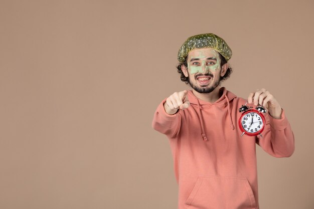 Vista frontal de los hombres jóvenes con máscara sosteniendo relojes sobre fondo marrón tiempo de terapia de spa cuidado corporal de la piel facial