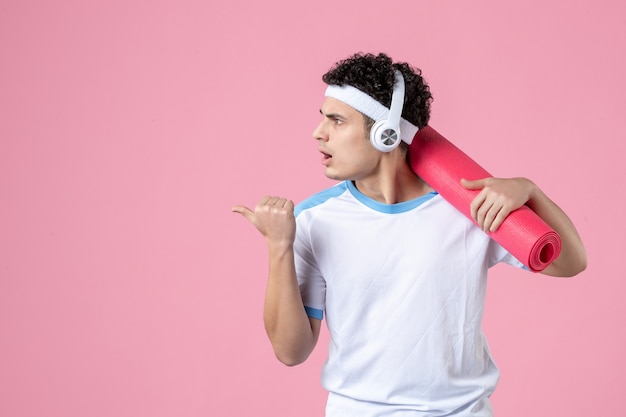 Vista frontal de los hombres jóvenes en ropa deportiva con estera de yoga y auriculares en la pared rosa