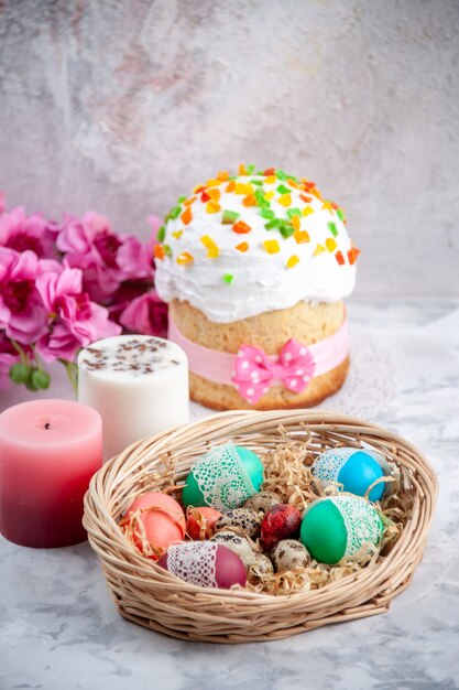 Vista frontal de los huevos de pascua de colores dentro de la canasta con velas y pastel sobre una superficie blanca
