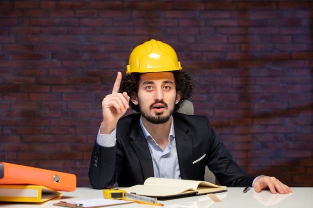 Vista frontal del ingeniero masculino sentado detrás del lugar de trabajo en traje y casco amarillo plan de negocio constructor trabajo corporativo contratista trabajo ocupación