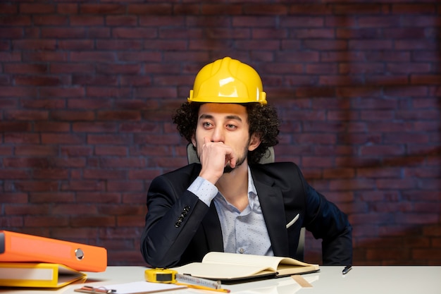 Vista frontal del ingeniero masculino sentado detrás del lugar de trabajo en traje y casco amarillo plan de negocio constructor trabajo corporativo proyecto de trabajo ocupación