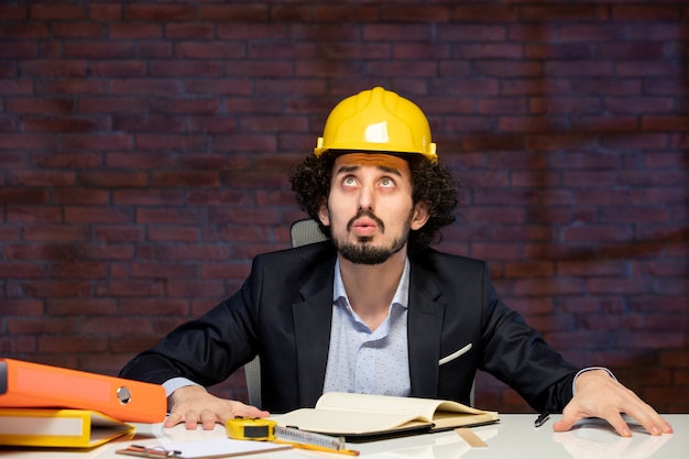 Vista frontal del ingeniero masculino sentado detrás del lugar de trabajo en traje y casco amarillo plan de proyecto empresarial contratista de trabajo corporativo trabajador constructor de ocupación