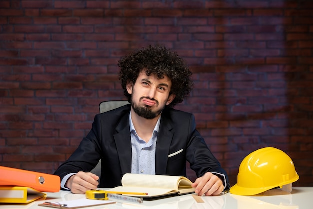 Vista frontal del ingeniero masculino sentado detrás del lugar de trabajo en traje contratista constructor de negocios trabajo trabajo corporativo en interiores