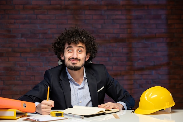 Vista frontal del ingeniero masculino sentado detrás del lugar de trabajo en traje contratista constructor trabajo corporativo negocio en el interior proyecto ocupación plan de trabajo