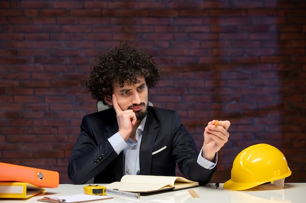 Vista frontal del ingeniero masculino sentado detrás del lugar de trabajo en traje plan de trabajo empresarial trabajo corporativo construcción constructores contratista proyecto