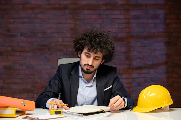 Vista frontal del ingeniero masculino sentado detrás del lugar de trabajo en traje y tomando notas trabajo corporativo de negocios contratista constructor de trabajo en el interior