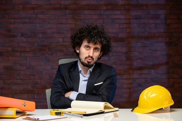 Vista frontal del ingeniero masculino sentado detrás del lugar de trabajo en traje de trabajo contratista en el interior del trabajo constructor corporativo