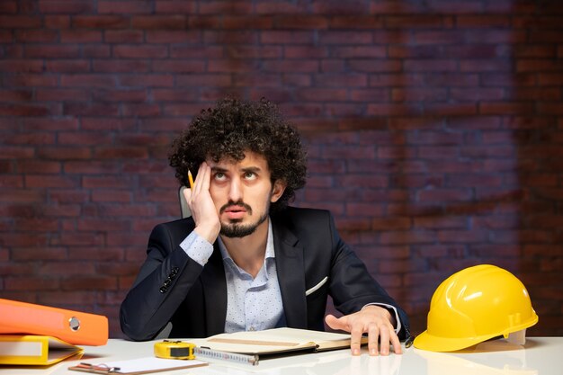 Vista frontal del ingeniero masculino sentado detrás del lugar de trabajo en traje de trabajo de contratista de negocios constructor corporativo agenda trabajo en interiores