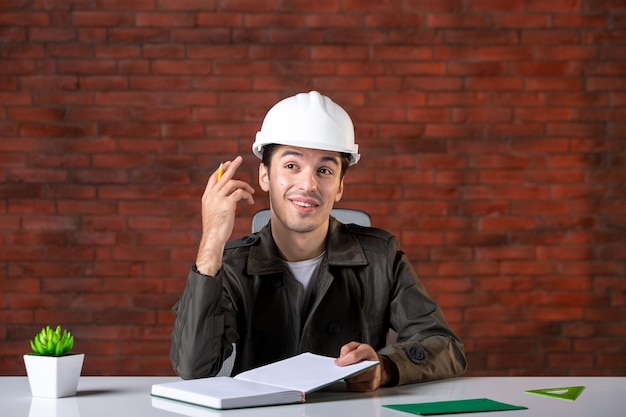 Vista frontal ingeniero masculino sentado detrás de su lugar de trabajo en el plan de documento de casco blanco contratista de negocios propiedad constructor de trabajo corporativo