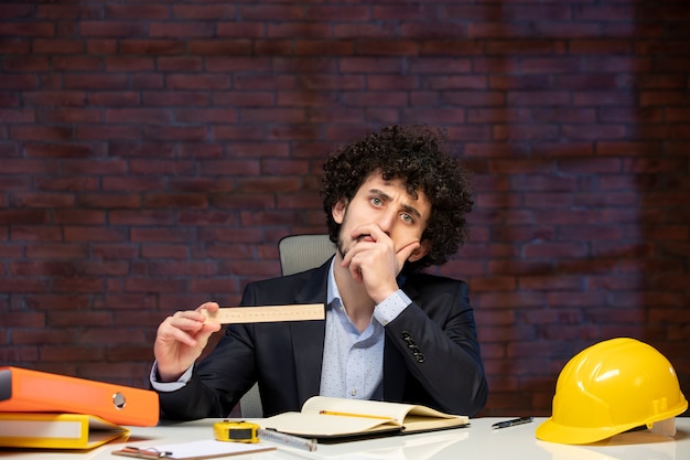 Vista frontal del ingeniero masculino sentado detrás de su lugar de trabajo en traje sosteniendo plan de regla trabajo de contratista trabajo de proyecto de construcción de ocupación corporativa