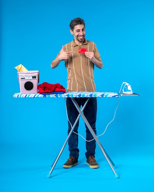 Foto vista frontal jovem do sexo masculino atrás da tábua de passar roupa segurando um cartão do banco vermelho sobre fundo azul dinheiro lavanderia trabalho doméstico máquina de lavar roupa cor ferro