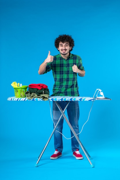 Foto vista frontal jovem do sexo masculino com tábua de engomar sorrindo sobre fundo azul roupas da casa máquina de lavar roupa limpa trabalho doméstico