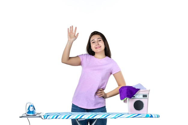 Foto vista frontal jovem dona de casa com roupas e tábua de engomar acenando e cumprimentando no fundo branco cor trabalho seco limpar mulher trabalho doméstico lavanderia emoção
