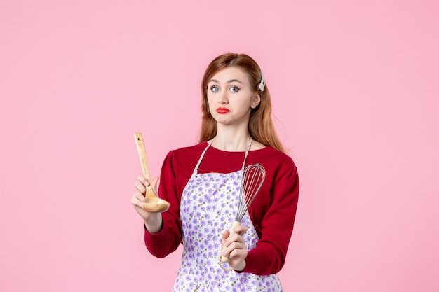vista frontal jovem dona de casa segurando um batedor e uma colher de pau no fundo rosa bolo cozinha profissão uniforme massa mulher cozinhar torta