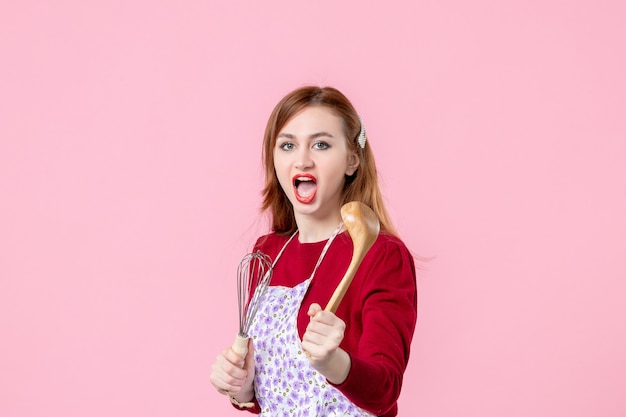 vista frontal jovem dona de casa segurando um batedor e uma colher de pau no fundo rosa cozinhando massa horizontal uniforme torta profissão mulher bolo cozinha