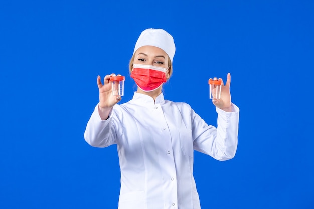vista frontal jovem enfermeira em traje médico com máscara vermelha segurando frascos sobre fundo azul Saúde vírus covid - hospital drogas médico medicina pandemia