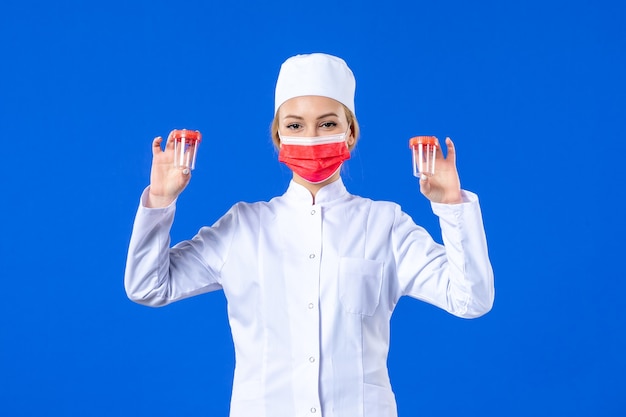 vista frontal jovem enfermeira em traje médico com máscara vermelha segurando frascos sobre fundo azul Saúde vírus pandemia covida- medicina hospital droga médico
