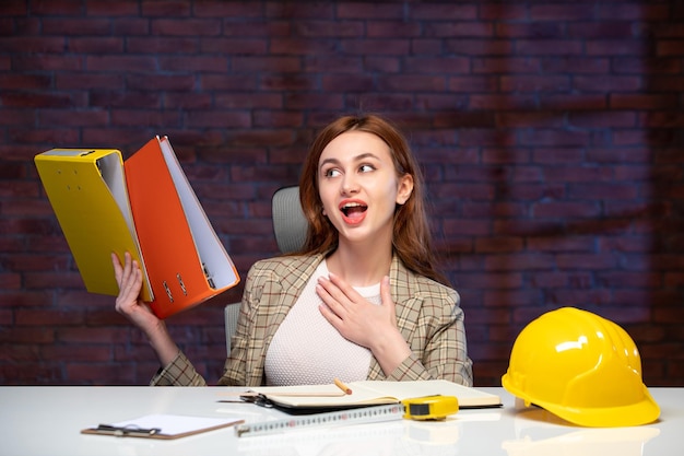 Foto vista frontal jovem engenheira sentada atrás de seu local de trabalho com documentos construtor de empregos agenda sucesso empresarial propriedade corporativa cores empreiteiro