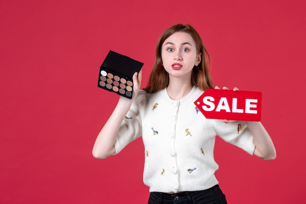 vista frontal jovem fêmea segurando caixa de pólvora e placa de identificação vermelha de venda em fundo vermelho compras mulher cor lábios marcha moda maquiagem beleza pele