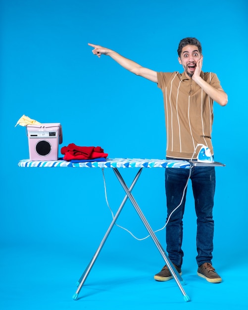 Vista frontal jovem homem atrás de tábua de passar roupa no fundo azul limpo emoção máquina de trabalho doméstico lavanderia homem lavando