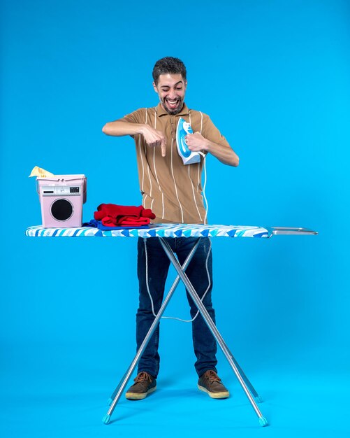 Foto vista frontal jovem homem atrás de tábua de passar roupa segurando ferro no fundo azul cor trabalho doméstico limpo máquina de lavar roupa homem emoção