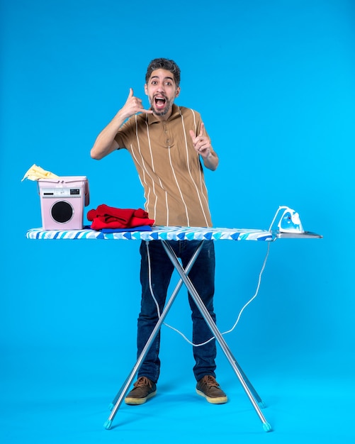 vista frontal jovem homem atrás de tábua de passar sobre o fundo azul cor trabalho doméstico lavanderia homem máquina de lavar roupa limpa emoção