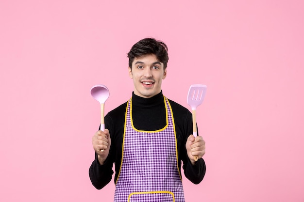 Vista frontal jovem macho na capa com colheres na cor de fundo rosa cozinhar comida profissão uniforme cozinha massa de trabalho