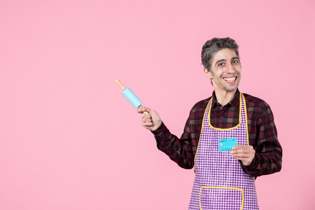 vista frontal jovem macho na capa segurando o rolo de massa e o cartão do banco no fundo rosa trabalho cozinhar marido trabalhador uniforme profissão dinheiro horizontal